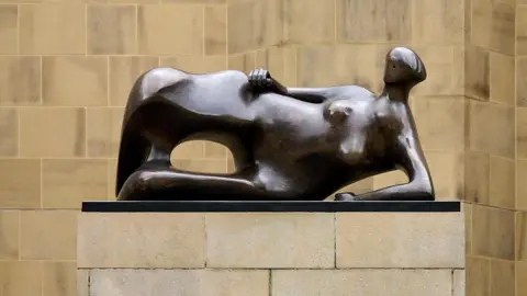 A bronze statue on a plinth. The figure is reclining in a relaxed manner. The wall behind is light stone.