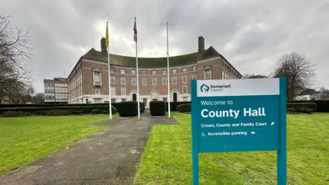 A sign pointing towards County Hall in Taunton, which is in the background. There are three flags, a green lawn and a path in front of the building. 