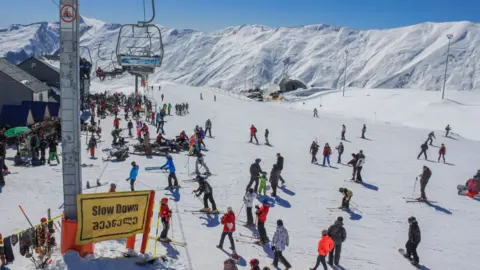 Skiers and snowboarders wait to join ski lifts at the Gudauri resort in Georgia. The ground is covered in snow and there are snow-capped mountains in the background