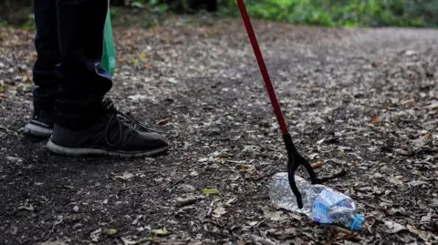 A generic photo of a person standing in a wooded area using a litter-picking tool to pick up a discarded water bottle. 