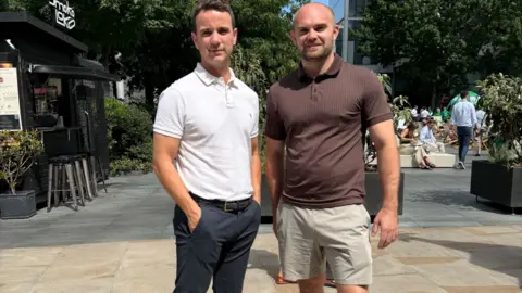 Two men stand in the street. One wears trousers, the other wears shorts