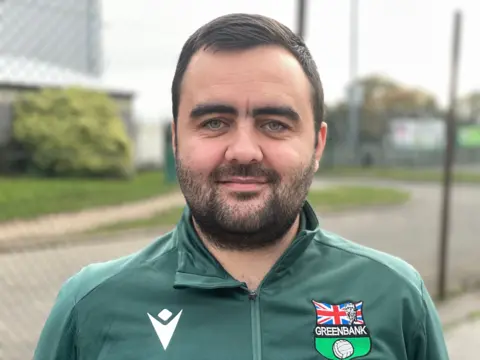 Mr Leeder wearing his Greenbank FC uniform in front of the closed pitches. 
He has a short beard and dark hair and his tracksuit is green with an emblem on his left chest.