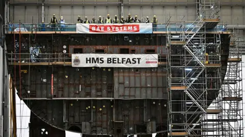 Getty Images A line of people wearing hi-vis vest and hard hats, look very small compared to the structure of the HMS Belfast ship they are standing on. The vessel is still in construction so lots of scaffolding is visible.