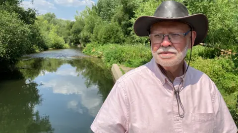 A man is wearing a hat and glasses, standing in front of a river.