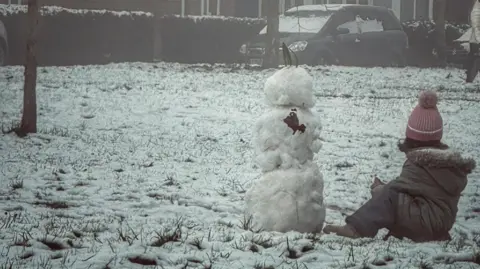 A young girl in a brown coat and waterproof trousers and pink hat sat on a snowy field next to a snowman. Houses can be seen in a street beyond.