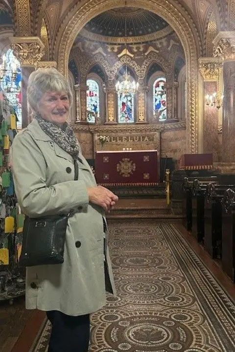 HANDOUT A smiling older woman stands in the aisle of a richly decorated chapel. She was wearing a light green coat. 