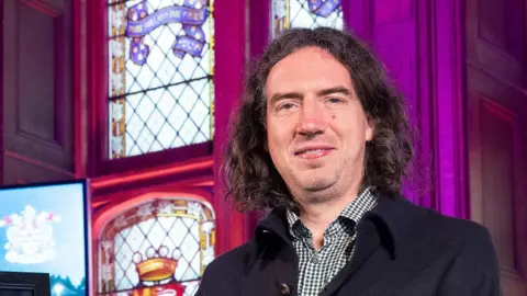 Gary Lightbody had dark shoulder length hair. He is wearing a black and white checked shirt over a navy coat, Behind him is a stained glass window. 