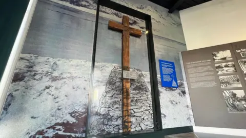 BBC A large wooden cross behind perspex in a black frame mounted on a wall in front of a picture of the Antarctic