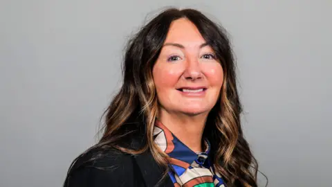 Redcar and Cleveland Borough Council A head-and-shoulders portrait of councillor Lynn Pallister. She has long dark soft curls with highlights. She is wearing a black blazer over a satin multicoloured patterned blouse. She is smiling at the camera. 