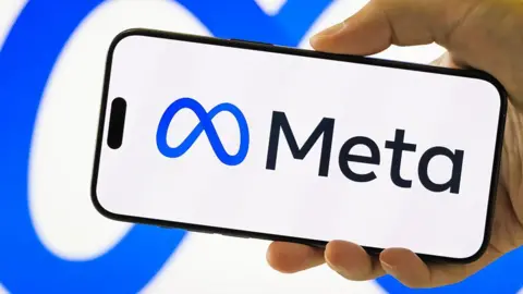 A picture of a hand holding a phone with the Meta logo and text in blue and black on a white background, behind it is a white background with the blue Meta logo on it.