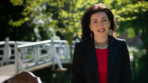 MCS Helen Pike has long dark hair and is smiling. She is stood outside, and is wearing a dark blazer over a red top.