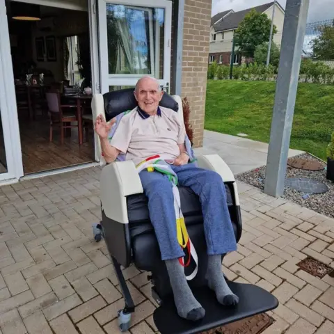 Jack, a smiling man sitting in a reclining wheelchair outside a care home, waving at the camera with the open patio doors and garden visible behind him.