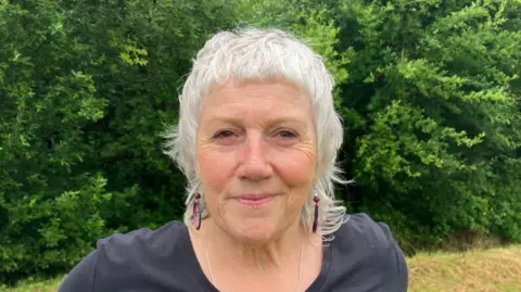 Diane Greer has long white hair and is wearing a black top and silver necklace.
She is standing in the festival grounds.