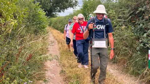 Andy Hockey John leads the way down a narrow country lane, with tall hedges either side. He holds a walking stick, with his family members walking in a line behind him. 