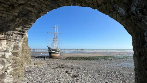 Julie Arthur A traditional tall ship sits upright on a sandy and stony beach, with the tide far out. It is a bright sunny day, with a cloudless blue sky. The view is framed through a stone arch, and there is a long iron pier in the distance. to the left.