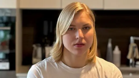 Headshot of Rūta Meilutytė, who is looking straight at the camera with a serious expression on her face. She has shoulder-length, straight blonde hair, with a long fringe. She wears a white round-necked t-shirt and is pictured in a kitchen.
