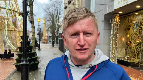 A man with blonde hair is standing outside in a town during the day. He is wearing a blue top over a grey hoodie. The street is adorned with various Christmas decorations and lights.