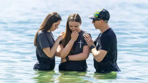Two young women and an older man stand waist deep in the sea. The man and one of the women are facing the woman in the centre and have their hands on her shoulders as she looks down towards the water.