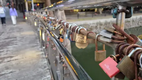 Alex Seabrook Locks attached to a metal footbridge. Some locks have names written on them. A body of water is visible beneath the bridge. Pedestrians and shops are visible at the far end of the bridge. 