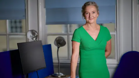 Developing health and independence Rosie Phillips stands at a desk in a green dress. Her hair is up and she is smiling. 