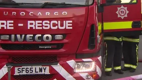 The front of a red fire engine that reads Fire - Rescue on the front in large white writing. The passenger side door is open and two fire fighters are talking next to the vehicle.