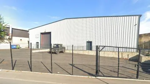 Google A warehouse, pictured from the outside behind a dark metal fence. A 4 x 4 is parked outside.