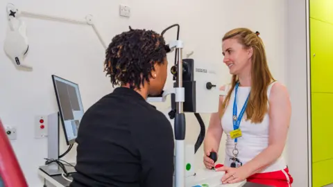 A woman with short hair is mid-way through an eye test being conducted by an optician with blonde and brown hair. The patient is resting her head on a machine and looking into a camera lens while the optician looks at the results on a monitor to her right. The room is white and another machine is leaning against the wall behind the patient.