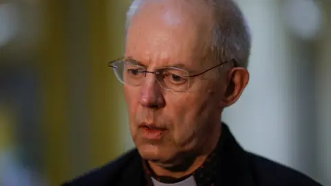 Reuters Justin Welby, wearing thin-framed spectacles, a dog collar and a black jacket, bears a serious expression as he speaks during a press interview