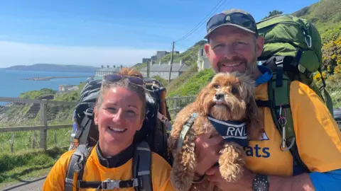 A couple in bright orange t-shirts that say RNLI lifeboats smile at the camera. Jason is holding their dog Teddy who is also wearing an RNLI bandana around his neck. The couple have huge backpacks on their backs and the coast path is behind them.