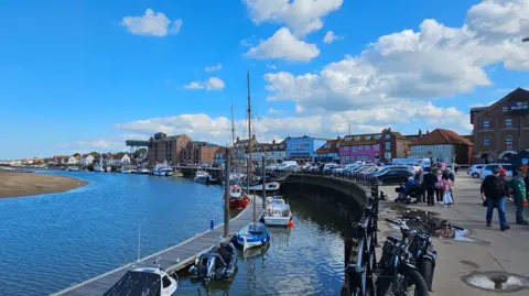 Paul Moseley/BBC A quayside showing a stretch of water, a curved harbour wall and a row of buildings beyond. Small boats are lined up by the quay wall and people are walking along a path to the right.