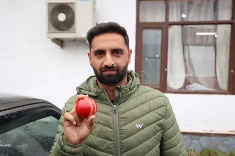 NurPhoto via Getty Images Auqib Nabi Dar poses with a red cricket ball in Baramulla, Jammu and Kashmir, India, on March 18, 2026. 