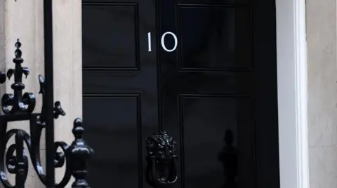 A black door in Downing Street with the Number 10 on it. A black railing can be seen in the foreground.