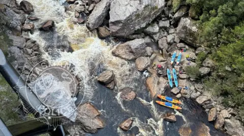 Tasmania Police Aerial photo shows part of the Franklin River in Tasmania. There are rocks in the river and six blue and orange kayaks are placed on the rocks. The river looks rapid and is surrounded by vegetation