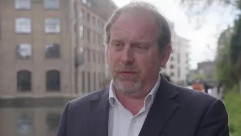 Jonathan Carr-West in a white shirt and blazer standing outside.