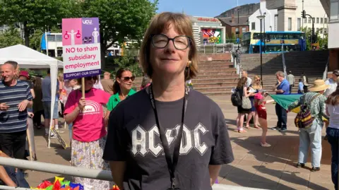 BBC/Naj Modak Annaliese Connolly wearing an AC/DC t-shirt and a staff ID badge stands in the foreground of an outdoor strike rally. In the background, people gather in a paved area, with one holding a pink UCU protest sign that reads "NO to sham subsidiary employment."