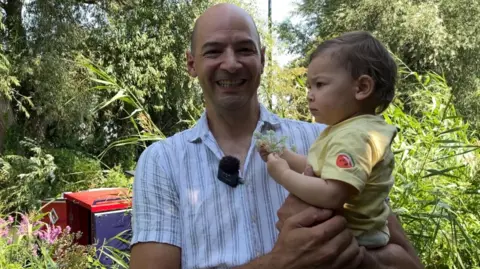 BBC Wayne has a bald head and is wearing a striped linen shirt. He is smiling whilst holding his young daughter. behind them is a red and blue canal boat.