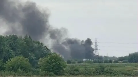 Maggie Riddell A distant view of thick black smoke billowing into the sky close to a pylon. There are fields and bushes in the foreground.