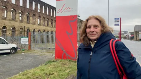 A woman with long, brown hair wearing a blue hooded coat standing next to a red and white sign spelling out Dalton Mills with a exterior Yorkshire stone walls of one of the mill buildings in the  background.  