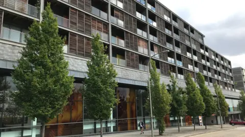 BBC/SAM READ A picture of part of the Vizion development. It is a large block of flats with what appears to be wooden cladding around balconies. The ground floor is a large shop. In front of the building is a line of green trees and a pavement. One person can be seen walking along the footpath.