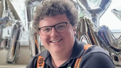 Charles Kinston, who has short brown curly hair and wears glasses, is smiling. Silver letter shaped balloons are in the background