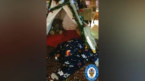 West Midlands Police A white fabric teepee style tent with green tinsel and fairy lights around it. There is a red cushion inside and a brown teddy bear. There is a sleeping bag inside which is navy blue and has astronauts, stars and rockets on it.