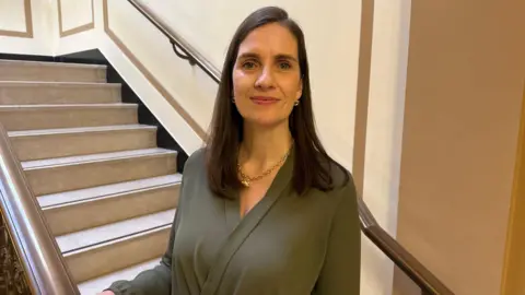 Sarah O'Toole is standing in a stairway. One of her hands is resting on the brown banister. Her long brown hair is straight. She's wearing gold earrings and a necklace. Her dress is dark green. 