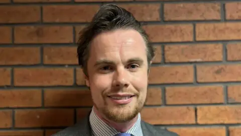 Councillor Philip Faircloth-Mutton is smiling at the camera he is wearing a suit and tie, with a striped shirt. He  is standing with against a brick wall 