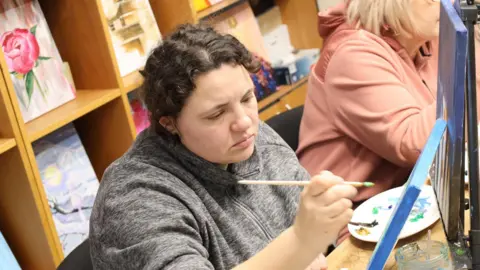 Veterans' House A woman with curly brown hair wearing a grey fleece paints.