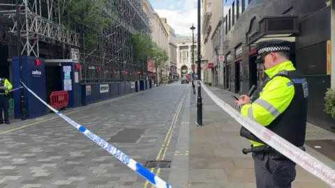 Police tape blocks off a central London street as officers stand on either side of the cordon near scaffolding and shopfronts, with the largely empty road stretching into the distance.