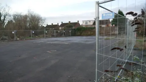 A derelict site surrounded by mental fencing. In the background there are three houses. The floor is covered in Tarmac.