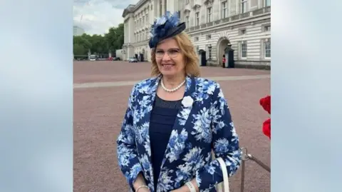 Supplied Jilly Crofts in front of Buckingham Palace in a blue, flowery suit jacket and hat