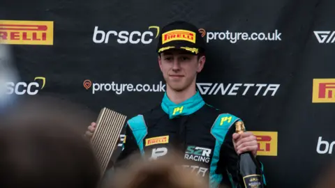 Jude Peters wearing a blue and black racing suit and a black cap with Pirelli written on it. He is holding a champagne bottle and a trophy.