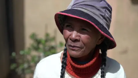 A head and shoulders shot of Neny Fara. She has a white top on over a red polo neck sweater. Two plaits of hair can be seen on either side of her face and they are emerging from a faded blue sun hat that is shielding her eyes.