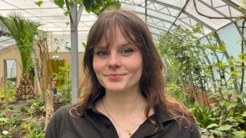 A woman brown hair smiling at the camera. She is wearing a brown polo top. Behind her are lots of plants. She is stood inside a large green house